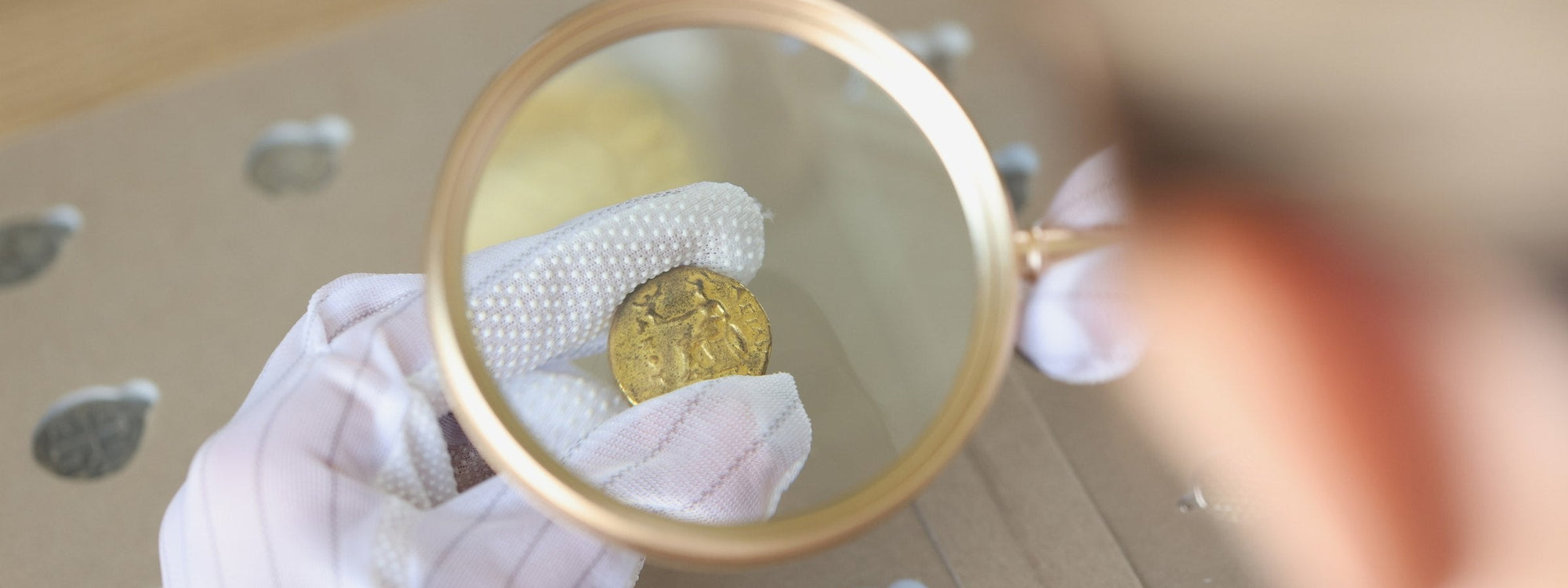 A gloved hand holding a magnifier over a historic gold coin on a coin tray